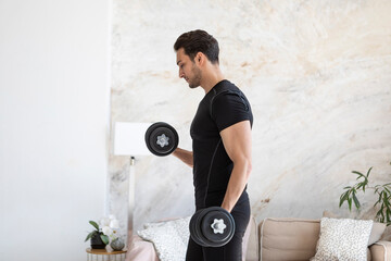 Side view of young handsome man in black sportswear doing biceps curl with dumbbells in living room at home. Healthy lifestyle and home workout concept - Powered by Adobe