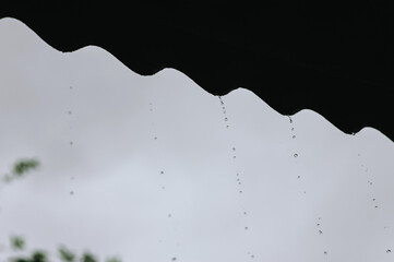 Drops of water from the rain drip from the roof of a slate house against the sky.