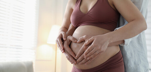 Pregnant young woman making heart with hands on belly and husband near her at home, closeup view with space for text. Banner design