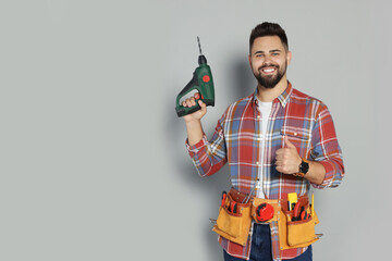 Young worker with power drill and tool belt on grey background. Space for text - obrazy, fototapety, plakaty