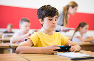 Fototapeta na wymiar Focused diligent tween schoolboy sitting at lesson in classroom, using mobile phone