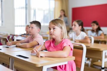Fototapeta premium Portrait of interested blonde tween girl studying in classroom, listening to schoolteacher and writing in notebook