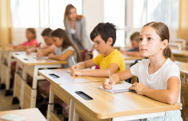Fototapeta premium Portrait of cute intelligent schoolgirl who writing exercises at lesson in primary school
