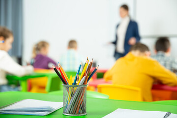 Closeup of multicolored pencils on school table on blurred background with teacher conducting lesson with children