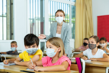 School boy and girl in face masks listening to teacher and writing exercises in notebook at lesson in primary school