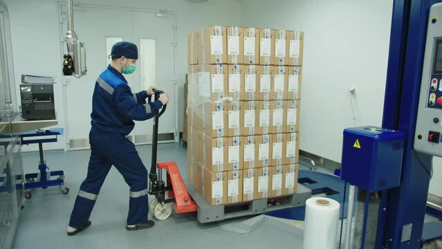 Loader in protective mask and uniform pushing cargo trolley manually at the pharmaceutical plant. factory worker transports the batch of packaged boxes of drugs to the weight machine.