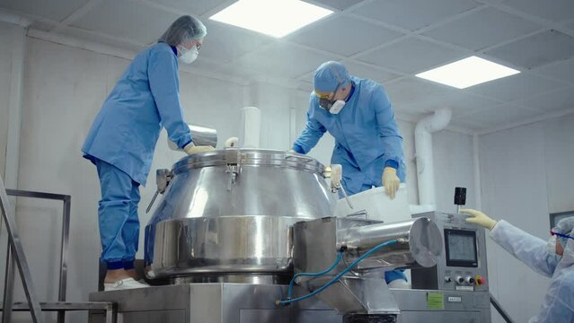 Pharmaceutical workers in protective masks and uniform work with medical equipment at medicine manufacturing factory. people pour the substance to automated machine for mixing and making drugs.
