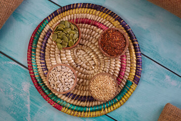 Sésamo, merkén, maravillas y semillas de zapallo en posillos de madera redondo sobre bandeja de mimbre de colores desde ángulo cenital.