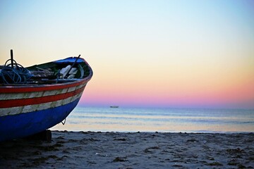 Fototapeta premium Boat with faded multi-color paint standing on the beach sand. In the background pink and blue light of dawn over the sea.