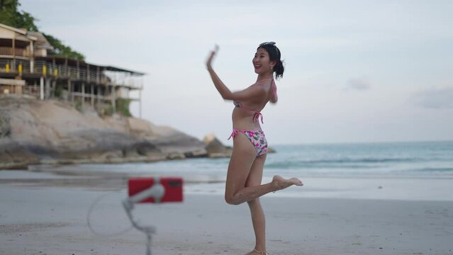Asian travel live streamer trying various poses to impress viewers of broadcast channel on sandy beach of Koh Phangan island in the evening