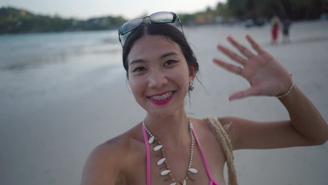 Happy asian blogger girl film selfie video wave hand saying hello standing on sandy beach  of tropical island in the evening