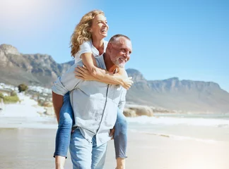 Fotobehang Happy mature couple enjoying vacation by the beach. Active senior husband giving his wife a piggyback ride while enjoying a sunny day outdoors. Energetic man and woman having fun while on holiday © Jade Maas/peopleimages.com
