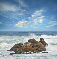 Copy space of a turbulent sea with rough tides and choppy waves from strong winds crashing onto big boulders at the beach with a cloudy blue sky background. Rocky coast in Western Cape, South Africa