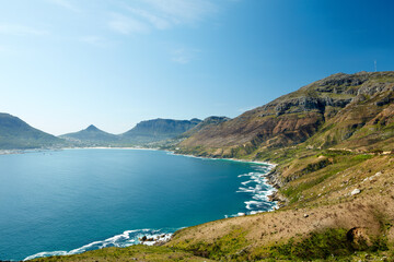 Landscape of rocky coastline near mountains in South Africa for a peaceful nature scene. Large blue lake or calm sea surrounded by vibrant green hills against a clear sky in Hout Bay.