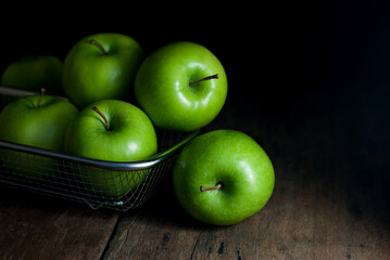 Delicious green apples are placed in a stainless steel basket and rest on a rustic wooden floor.