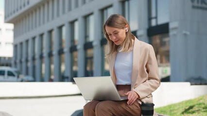 Smiling business woman talking to colleagues on laptop while sitting near office building on bench. Happy female manager in wireless earphones making video call conference outdoors. - Powered by Adobe