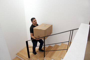 Latino adult man carries a heavy cardboard box up stairs which causes severe pain in his lower back and back
