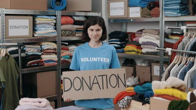 Portrait of energetic caucasian woman looking at the camera and smiling while holding banner with donation word in charity shop. Volunteer working in organization and sorting donated clothes