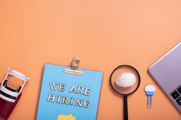 Wooden letters with phrase we are hiring on clip-board with magnifying glass, braine, stamp and laptop top view. Search worker concept