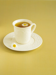 Сhamomile tea in a white cup on a white saucer. Сhamomile flower on tea surface. Yellow background