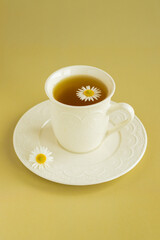 Сhamomile tea in a white cup on a white saucer. Сhamomile flower on tea surface. Yellow background