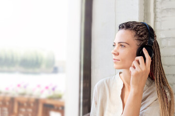 young woman with afro-braids listens to music or an audio book in headphones near the window,relaxation, Listen to an audio book, Alone with yourself,music enjoyment concept