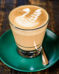 A latte foam art on a cup of coffee, placed in a drinking glass and green saucer with a wood grain background