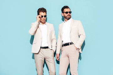 Portrait of two handsome confident stylish hipster lambersexual models. Sexy modern men dressed in white elegant suit. Fashion male posing in studio near blue wall. In sunglasses