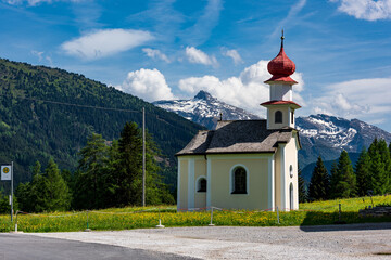 Eine kleine Kapelle auf einer grünen Wiese am Straßenrand einer Landstraße in den Alpen im Sommer