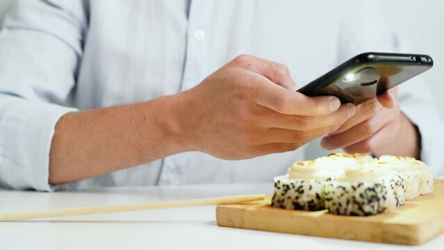 Close-up of man in shirt taking photo of traditional japanese sushi food on mobile phone for posting on social networks. Selective focus on smartphone and male hands.