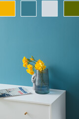 Vase with yellow flowers and magazine on table near blue wall in room. Different color patterns
