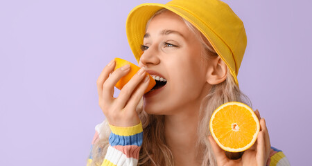 Beautiful young blonde woman eating orange on lilac background