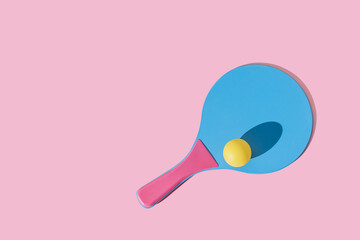 Single blue and pink colourful beach tennis paddle racket with a yellow ping pong ball against pink background. Flat lay pattern with repetition .Outdoors summer activity, fun relax sport idea. - obrazy, fototapety, plakaty