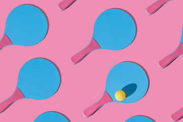 Many blue and pink colourful beach tennis paddle racket with a yellow ping pong ball against pink background. Flat lay pattern with repetition .Outdoors summer activity, fun relax sport idea.