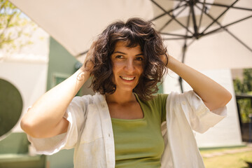 Fototapeta premium Joyful young caucasian girl smiling looking at camera ruffling her hair standing outdoors. Brunette woman is resting on weekends in summer. Lifestyle, different emotions, leisure concept.