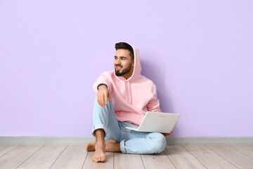 Handsome barefoot man with laptop sitting near lilac wall