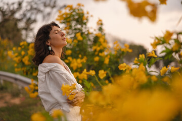 Cute young caucasian woman breathes fresh air with her eyes closed near flowering plants. Brunette...