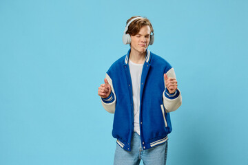 Fototapeta premium naughty handsome guy student stands dancing with hand movements on a light blue background listening to music through headphones