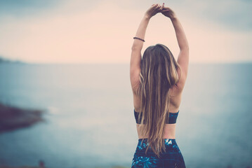 Back view of healthy fit body young woman with long beautiful brown hair stretching arms after workout and enjoying sea view and serene nature in front of her. Concept of active people lifestyle