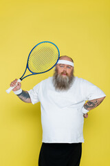 bearded plus size tennis player standing with hand on waist and looking at camera isolated on yellow.