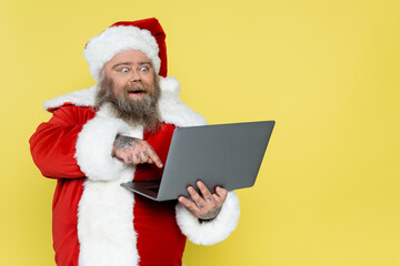 amazed overweight santa claus pointing at laptop isolated on yellow.