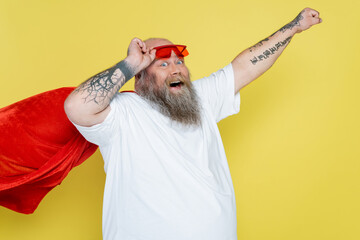 tattooed plus size man in superhero cloak posing with outstretched hand isolated on yellow.