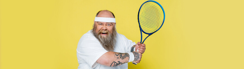 thrilled plus size man with beard and tennis racquet looking at camera isolated on yellow, banner.