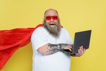 thrilled overweight man in red sunglasses and superhero cloak using laptop isolated on yellow.