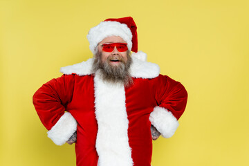 cheerful overweight santa claus in red sunglasses standing akimbo isolated on yellow.