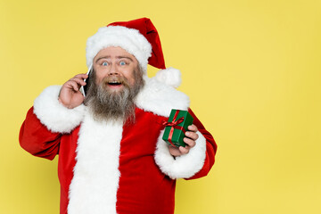 astonished plus size santa claus showing gift box while talking on smartphone isolated on yellow.