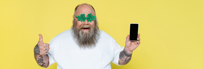 excited plus size man in clover-shaped glasses holding smartphone and showing thumb up isolated on yellow, banner.