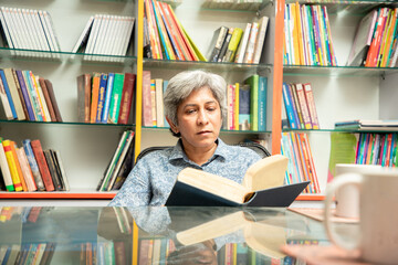 Senior indian woman reading a book at library sitting with bookshelf behind her, knowledge and...