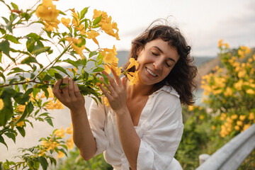 Positive young caucasian woman enjoying scent of flowers closing her eyes outdoors. Brunette wears white shirt on casual day. Relaxation concept