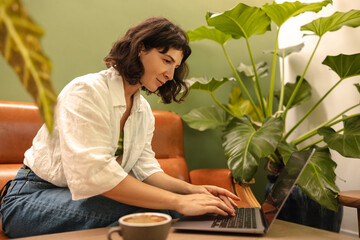 Busy young caucasian woman sits in cafe, drinks coffee and works on laptop in spare time. Brunette with wavy hair wears shirt and jeans. Technology concept